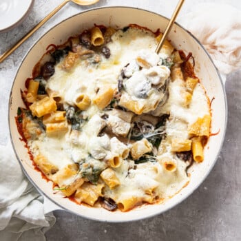 A baked pasta dish with rigatoni, melted cheese, spinach, and mushrooms in a round white pan. A gold serving spoon and fork are in the pan, with a napkin and bowl nearby on a light surface.