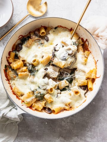 A baked pasta dish with rigatoni, melted cheese, spinach, and mushrooms in a round white pan. A gold serving spoon and fork are in the pan, with a napkin and bowl nearby on a light surface.