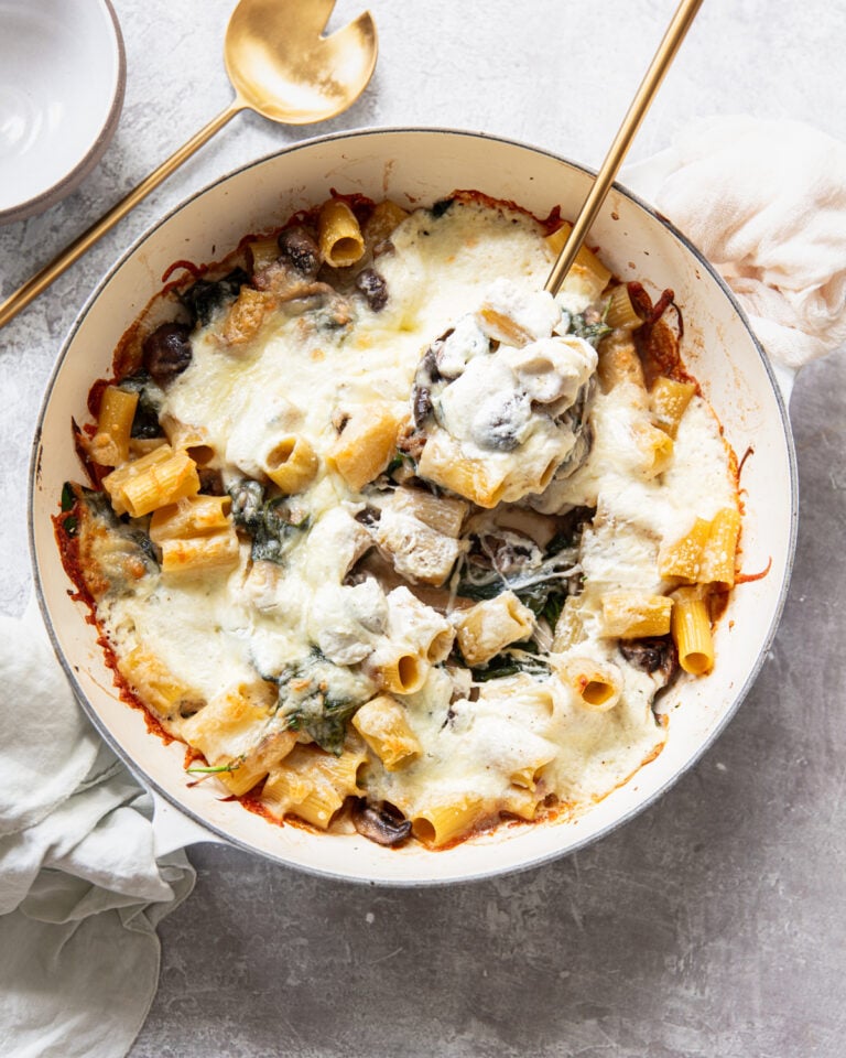 A baked pasta dish with rigatoni, melted cheese, spinach, and mushrooms in a round white pan. A gold serving spoon and fork are in the pan, with a napkin and bowl nearby on a light surface.