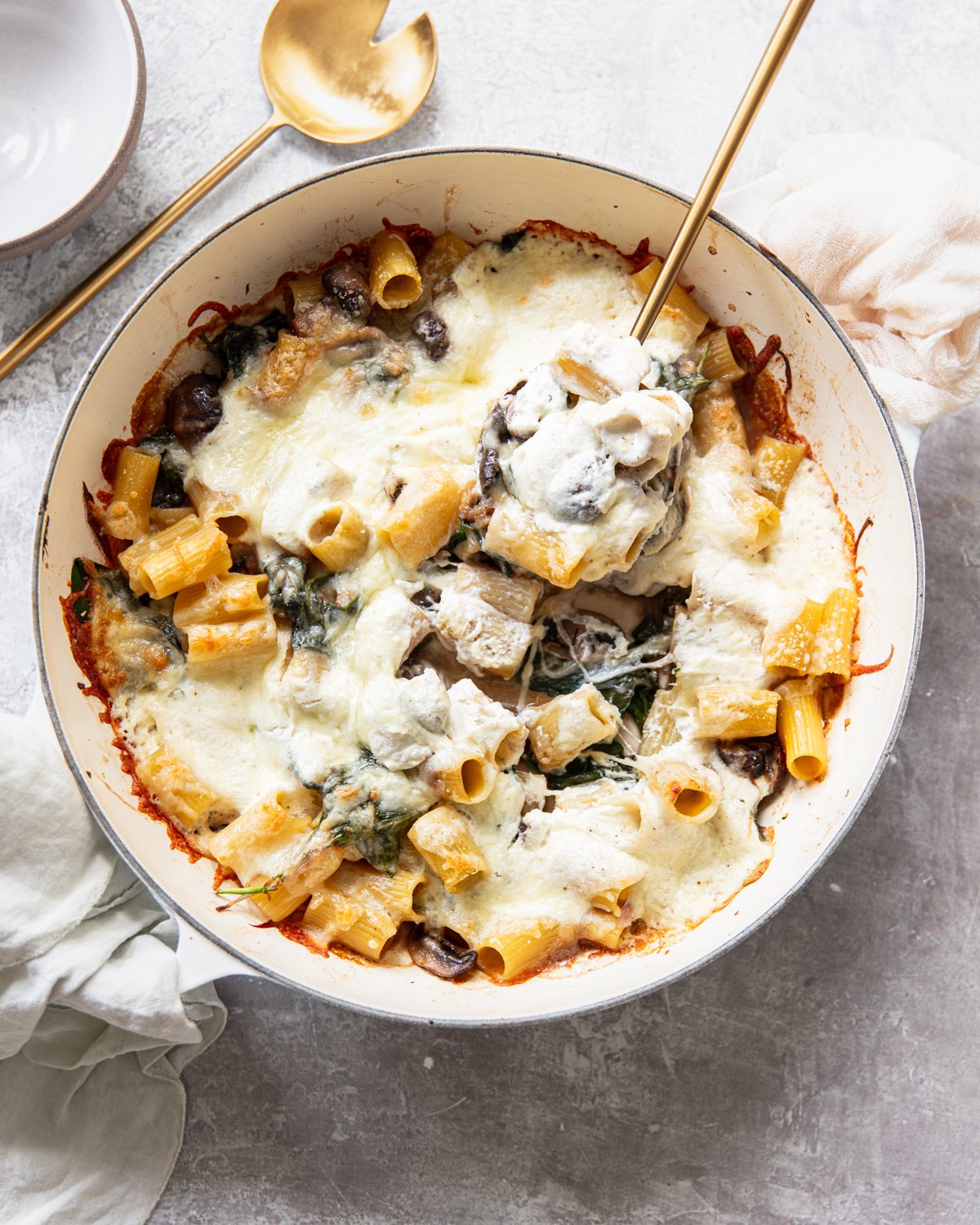 A baked pasta dish with rigatoni, melted cheese, spinach, and mushrooms in a round white pan. A gold serving spoon and fork are in the pan, with a napkin and bowl nearby on a light surface.