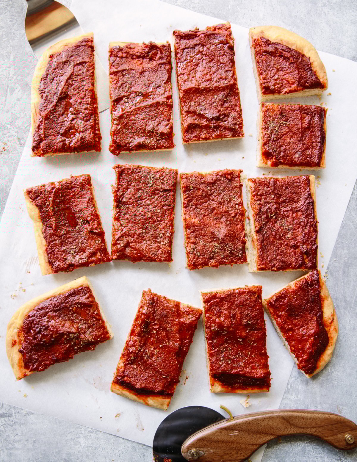 Rectangular slices of thick, square pizza with a layer of tomato sauce and seasonings on top, arranged on parchment paper with a pizza cutter nearby.
