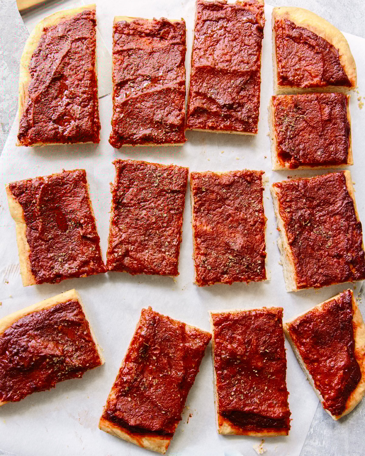 Rectangular slices of pizza with a thick layer of tomato sauce and herbs on top, arranged on parchment paper. The pizza has no visible cheese or other toppings.