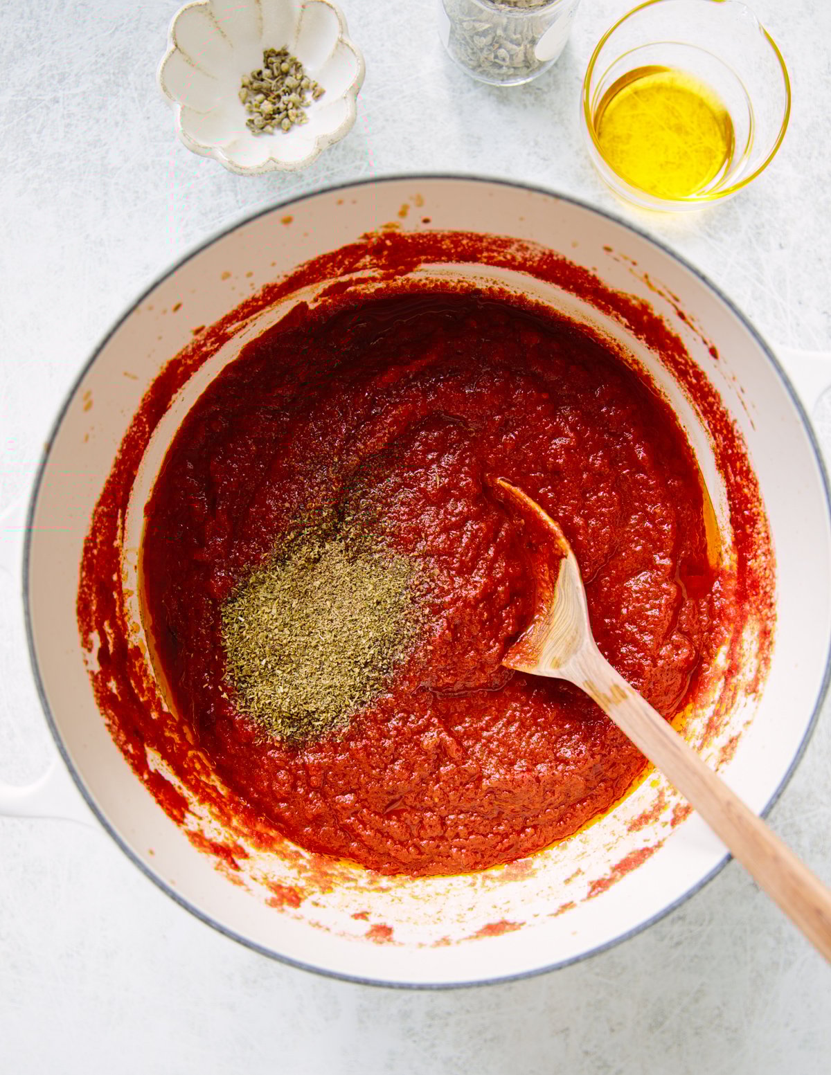 A large white pot filled with red tomato sauce being stirred by a wooden spoon. Dried herbs are sprinkled on top. Nearby are a bowl of peppercorns and a glass container of olive oil.