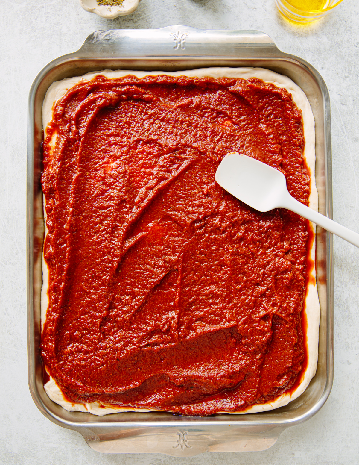 A rectangular baking pan with pizza dough topped with tomato sauce being spread evenly with a white spatula. Olive oil and a spice shaker are visible in the background.