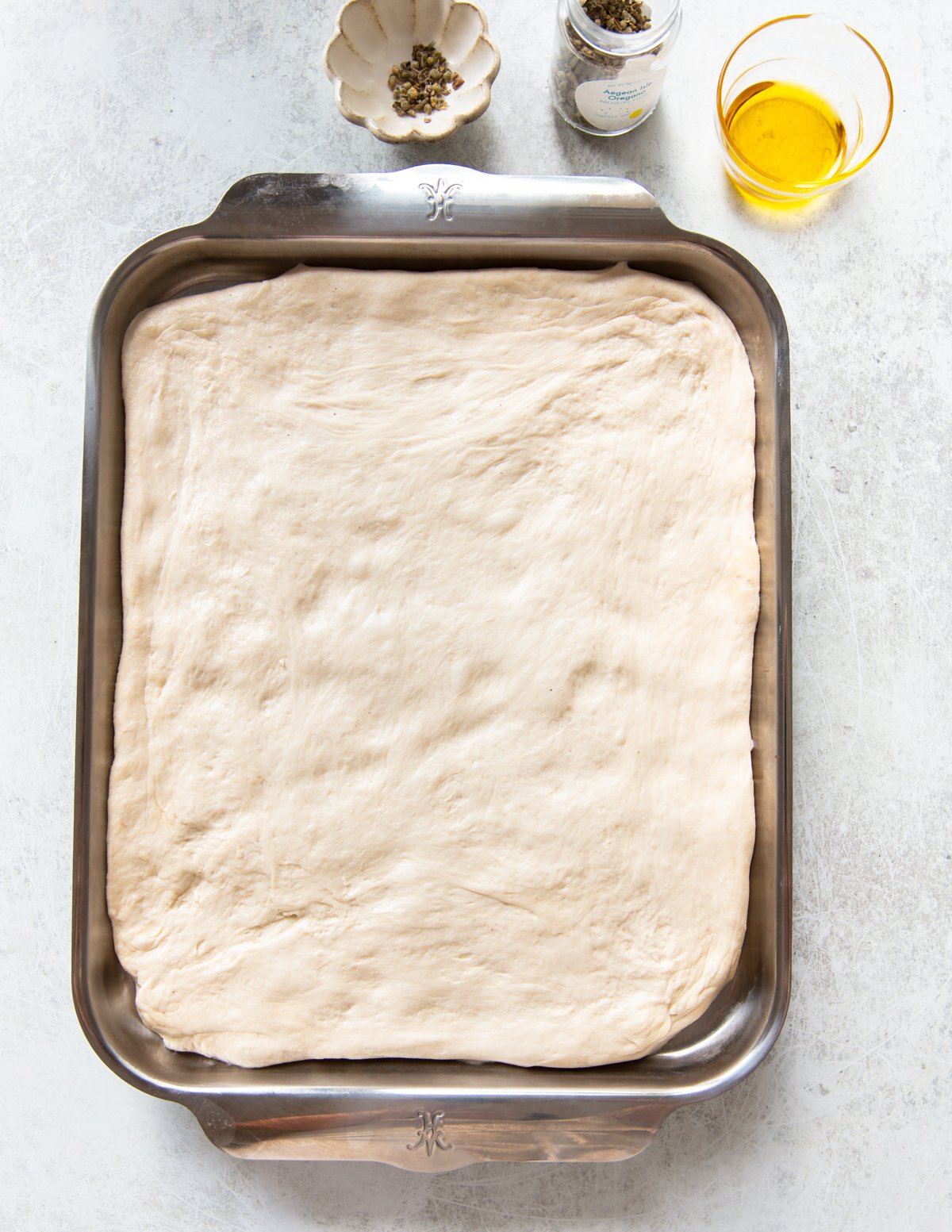 A rectangular baking tray filled with uncooked pizza dough. Nearby are a small bowl of pepper, a jar of sea salt, and a glass bowl of olive oil on a light-colored surface.