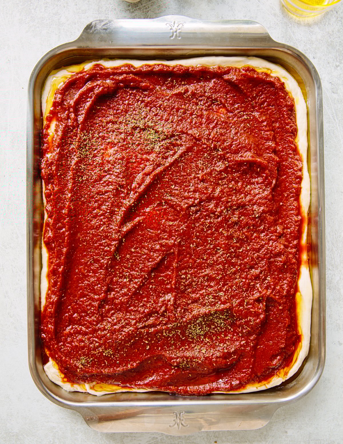 A rectangular baking pan filled with pizza dough topped with a thick layer of tomato sauce and sprinkled with dried oregano, ready to be baked.