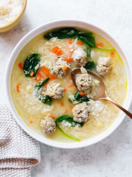 A bowl of Italian wedding soup with meatballs, orzo pasta, spinach, and diced carrots, served with a gold spoon. A small bowl of grated cheese and a textured napkin are beside the soup on a light surface.