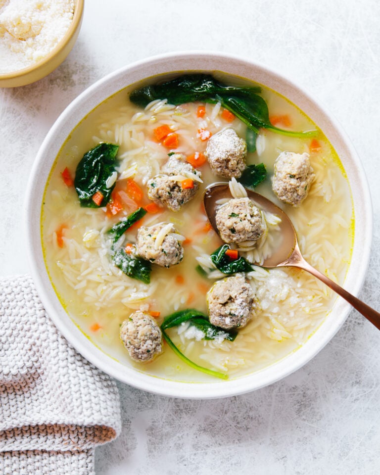 A bowl of Italian wedding soup with meatballs, orzo pasta, spinach, and diced carrots, served with a gold spoon. A small bowl of grated cheese and a textured napkin are beside the soup on a light surface.
