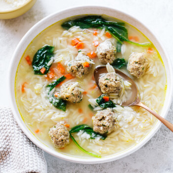 A bowl of soup with small meatballs, orzo pasta, spinach, and diced carrots, with a spoon resting inside. A knitted cloth and a bowl of grated cheese are nearby on a light surface.