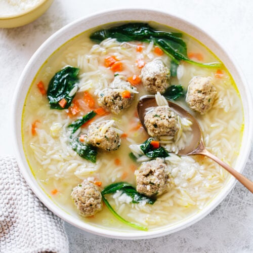 A bowl of soup with small meatballs, orzo pasta, spinach, and diced carrots, with a spoon resting inside. A knitted cloth and a bowl of grated cheese are nearby on a light surface.