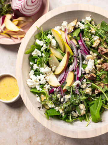 A bowl of salad with arugula, sliced pears, red onion, walnuts, and crumbled cheese, next to a small dish of dressing and a plate with sliced pear and red onion.