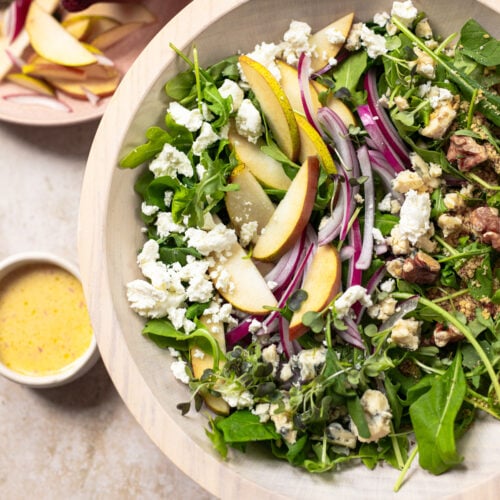 A bowl of salad with arugula, sliced pears, red onion, walnuts, and crumbled cheese, next to a small dish of dressing and a plate with sliced pear and red onion.