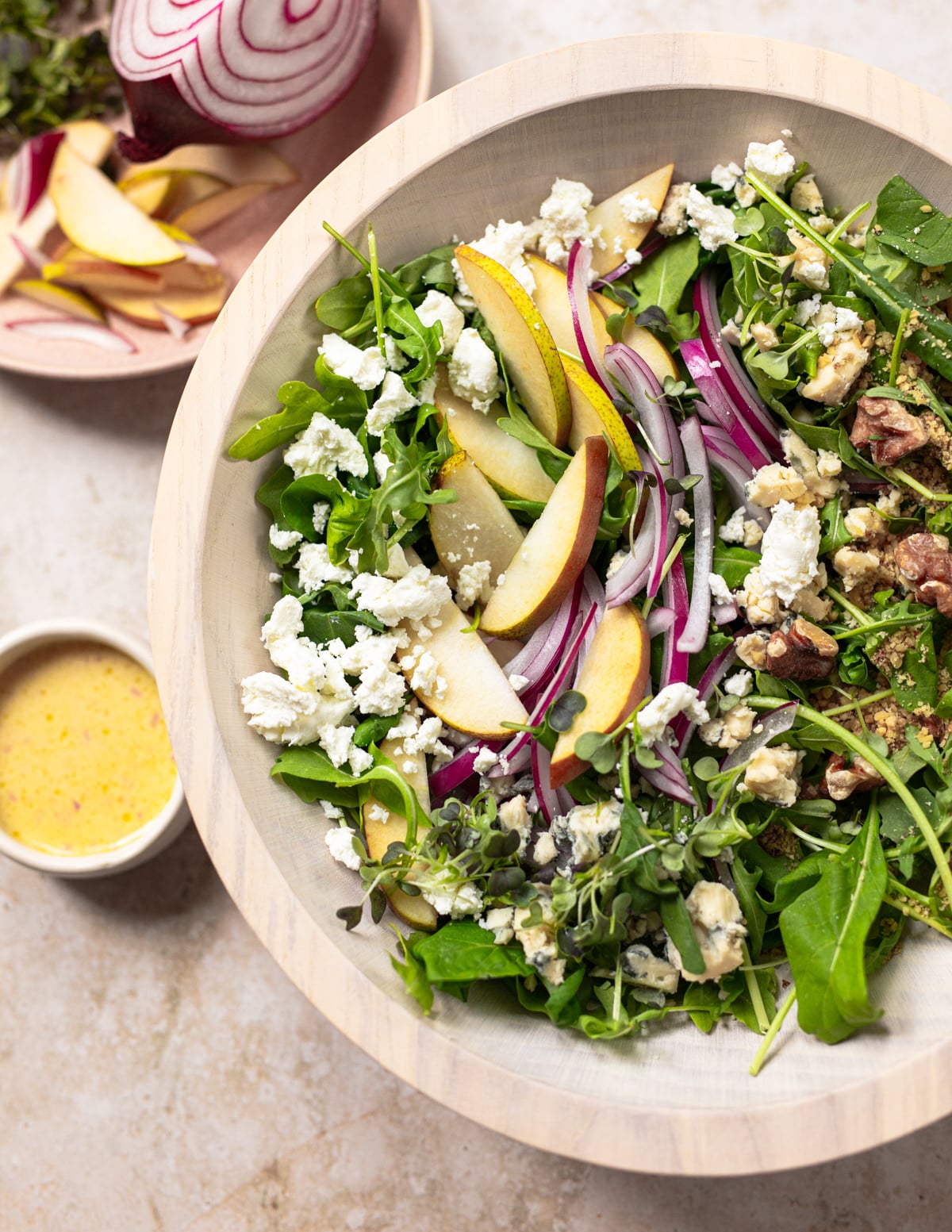 A bowl of salad with arugula, sliced pears, red onion, walnuts, and crumbled cheese, next to a small dish of dressing and a plate with sliced pear and red onion.