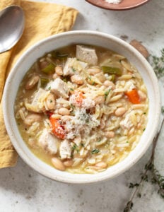 A bowl of chicken orzo soup with chunks of chicken, carrots, celery, white beans, and orzo pasta, garnished with grated cheese. A spoon and yellow napkin are beside the bowl on a light surface.