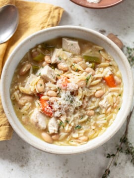 A bowl of chicken orzo soup with chunks of chicken, carrots, celery, white beans, and orzo pasta, garnished with grated cheese. A spoon and yellow napkin are beside the bowl on a light surface.