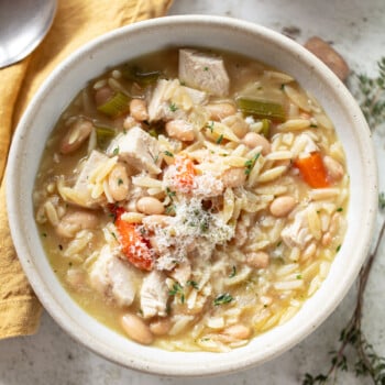 A bowl of chicken orzo soup with chunks of chicken, carrots, celery, white beans, and orzo pasta, garnished with grated cheese. A spoon and yellow napkin are beside the bowl on a light surface.