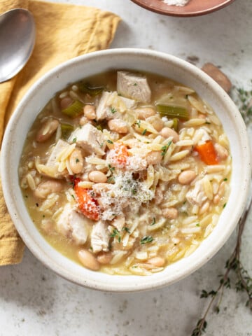 A bowl of chicken orzo soup with chunks of chicken, carrots, celery, white beans, and orzo pasta, garnished with grated cheese. A spoon and yellow napkin are beside the bowl on a light surface.