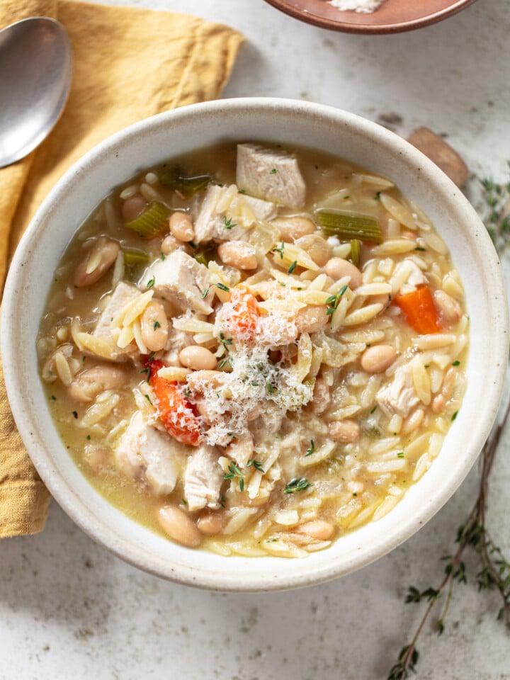 A bowl of chicken orzo soup with chunks of chicken, carrots, celery, white beans, and orzo pasta, garnished with grated cheese. A spoon and yellow napkin are beside the bowl on a light surface.