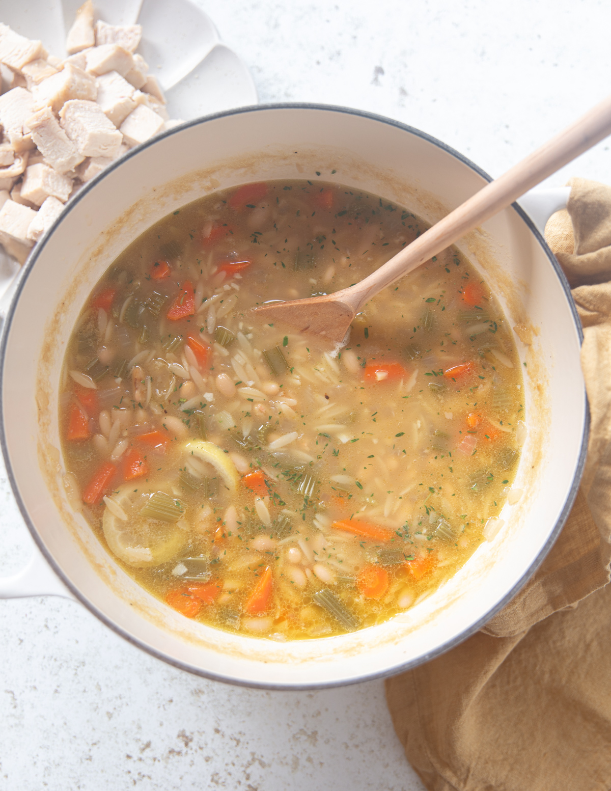 A pot of soup with orzo pasta, chopped carrots, celery, and herbs, stirred with a wooden spoon. A bowl of diced cooked chicken sits nearby on the counter.