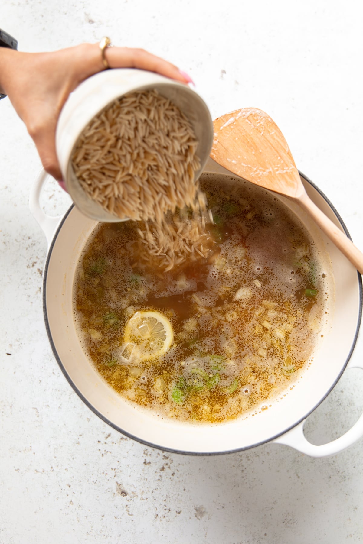 A hand pours dry orzo pasta from a cup into a white pot of simmering soup with herbs, vegetables, and a slice of lemon; a wooden spoon rests on the pots edge.
