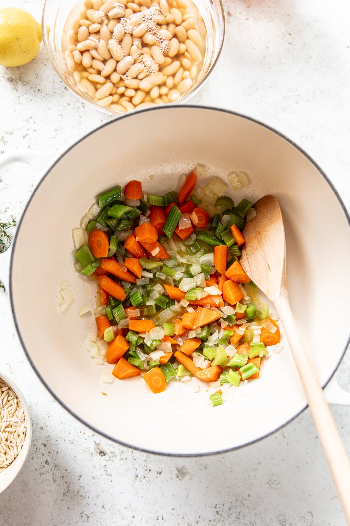 A white pot with chopped carrots, celery, and onions being sautéed with a wooden spoon. Nearby are bowls of white beans, uncooked rice, and half a lemon on a light countertop.