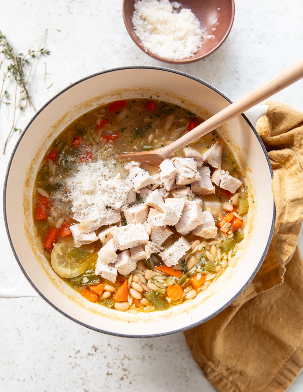 A pot of chicken and vegetable soup, topped with grated cheese and chunks of cooked chicken, sits on a white surface with a wooden spoon. A bowl of grated cheese and a yellow cloth are nearby.