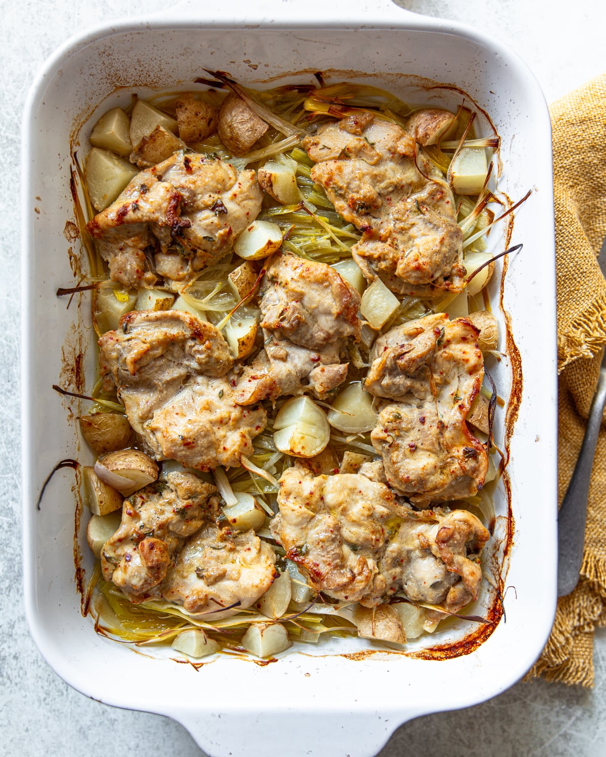 A white baking dish filled with baked chicken thighs on a bed of sliced potatoes, shallots, and leeks, showing golden, roasted edges and small browned spots, with a yellow kitchen towel beside the dish.