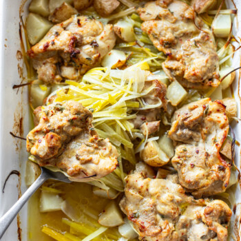 Baked chicken thighs with a golden-brown crust, surrounded by diced potatoes and sliced leeks in a white roasting dish. A serving spoon lifts one piece, displaying the tender chicken and vegetables.