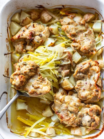 Baked chicken thighs with a golden-brown crust, surrounded by diced potatoes and sliced leeks in a white roasting dish. A serving spoon lifts one piece, displaying the tender chicken and vegetables.