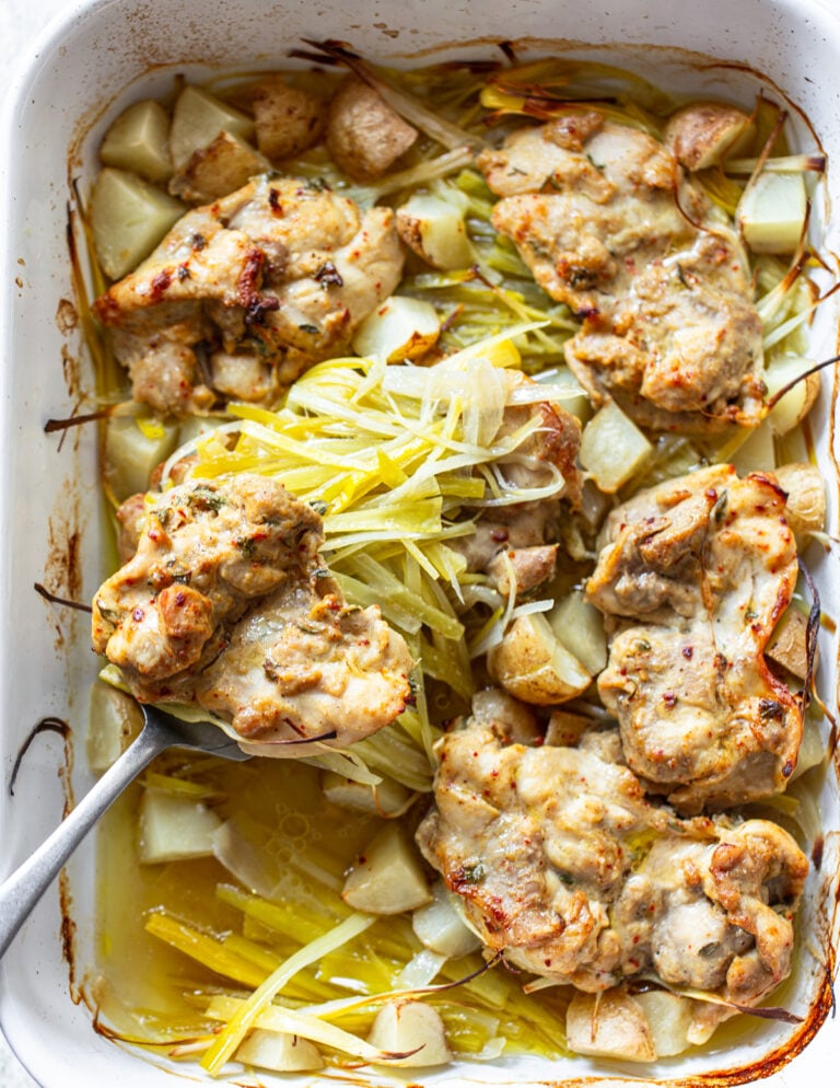 Baked chicken thighs with a golden-brown crust, surrounded by diced potatoes and sliced leeks in a white roasting dish. A serving spoon lifts one piece, displaying the tender chicken and vegetables.