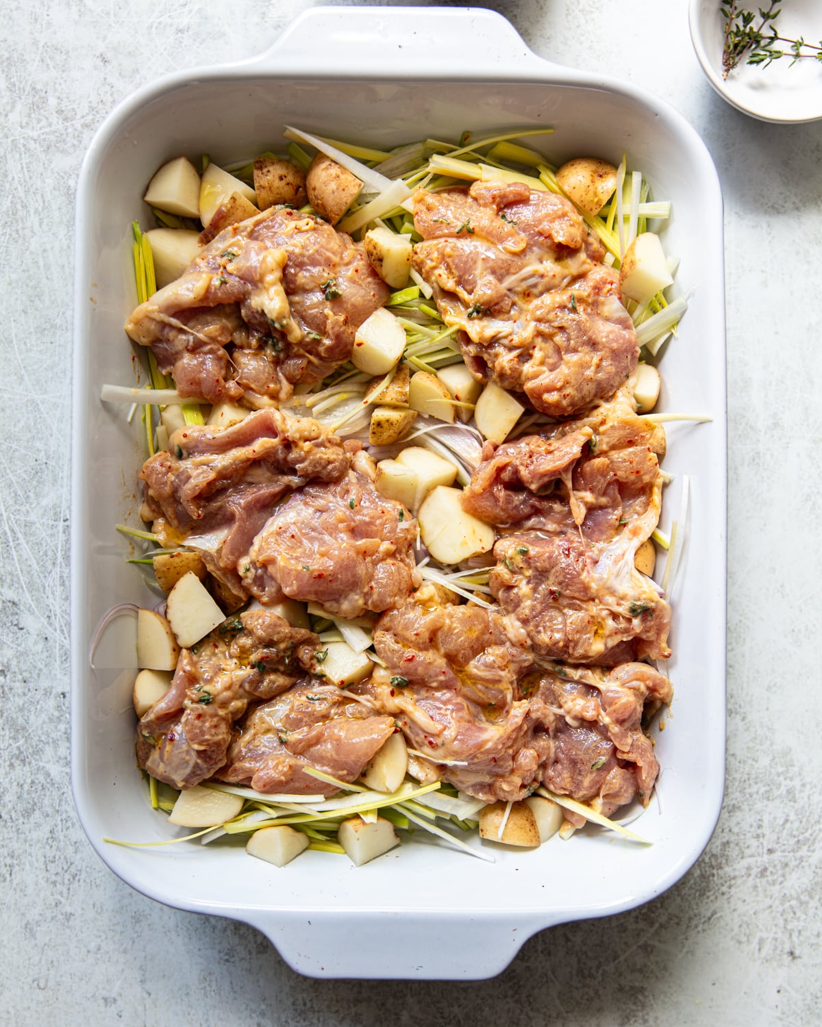 Raw seasoned chicken thighs arranged in a white baking dish, placed over a bed of chopped potatoes, leeks, ready for roasting. A small bowl with herbs is visible in the corner.