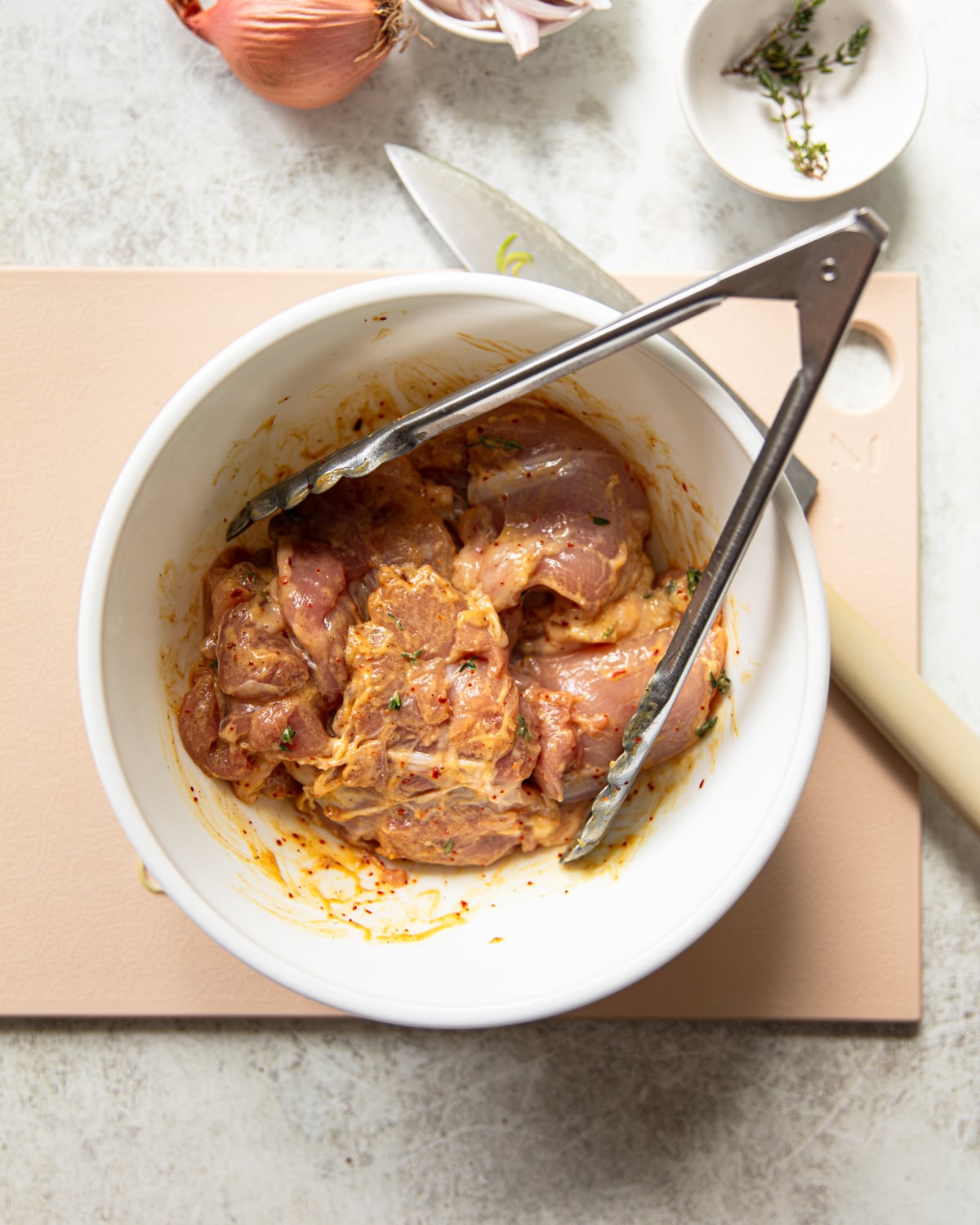 A white bowl with mustard and herb marinated raw chicken and metal tongs sits on a cutting board. Nearby are a knife, a shallot, and a small dish with thyme. The background is a light countertop.