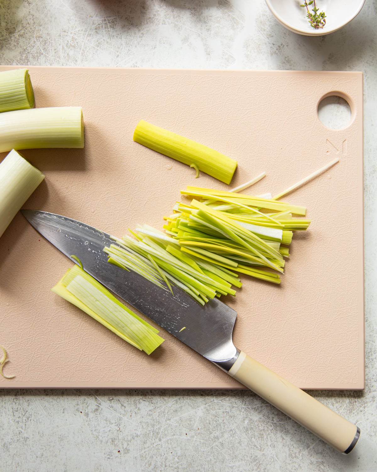A knife rests on a light cutting board, surrounded by chopped and whole pieces of leek. Some leek strips are thinly sliced, while others remain in larger sections.
