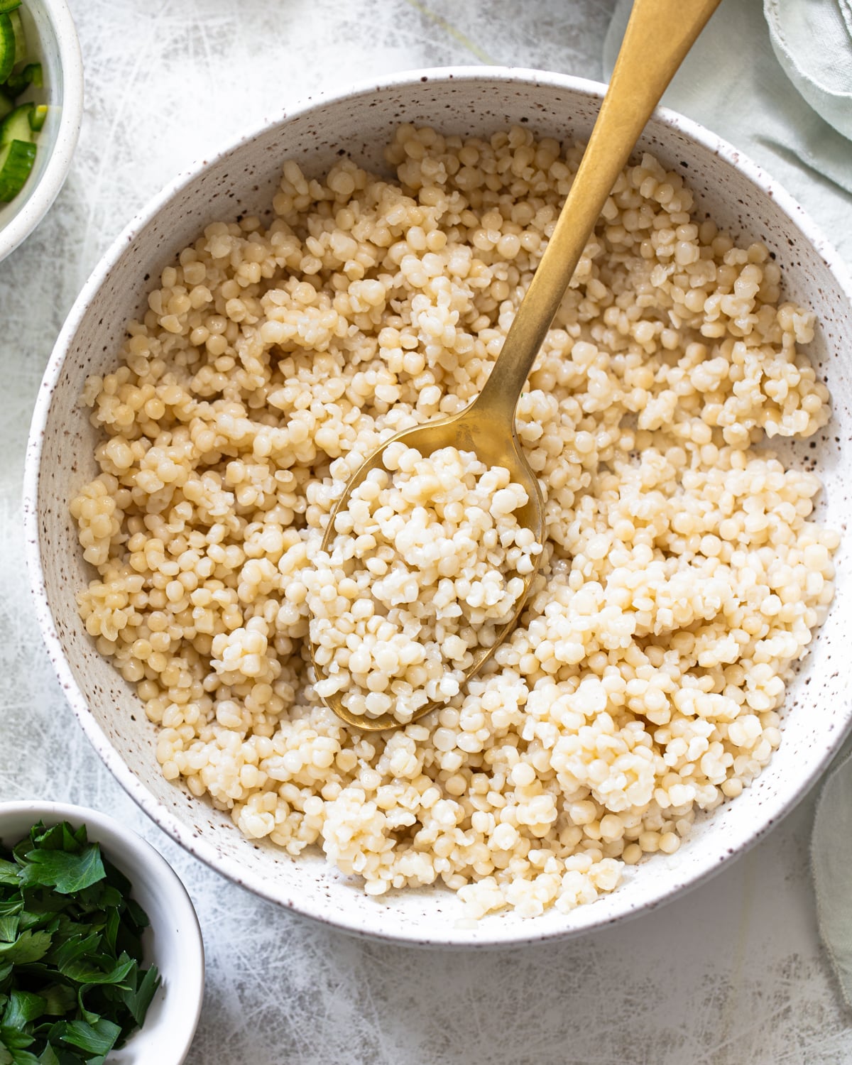 A speckled bowl filled with cooked pearl couscous, with a gold spoon resting inside. Fresh chopped herbs sit in a small bowl nearby on a light-colored surface.