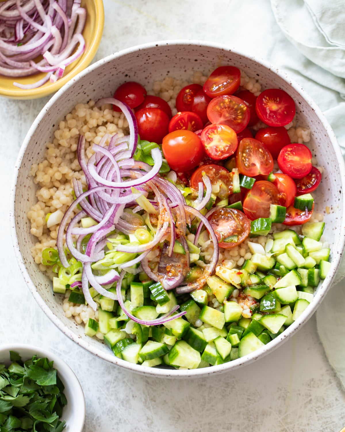 A bowl filled with couscous, sliced red onions, halved cherry tomatoes, and chopped cucumbers, with a small bowl of chopped herbs and more sliced onions nearby.