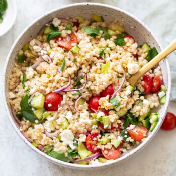 A bowl of couscous salad with cherry tomatoes, sliced red onions, cucumber, feta cheese, and fresh parsley, served with a wooden spoon on a light-colored table.