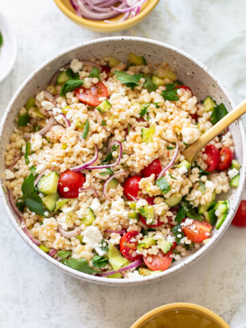 A bowl of couscous salad with cherry tomatoes, sliced red onions, cucumber, feta cheese, and fresh parsley, served with a wooden spoon on a light-colored table.