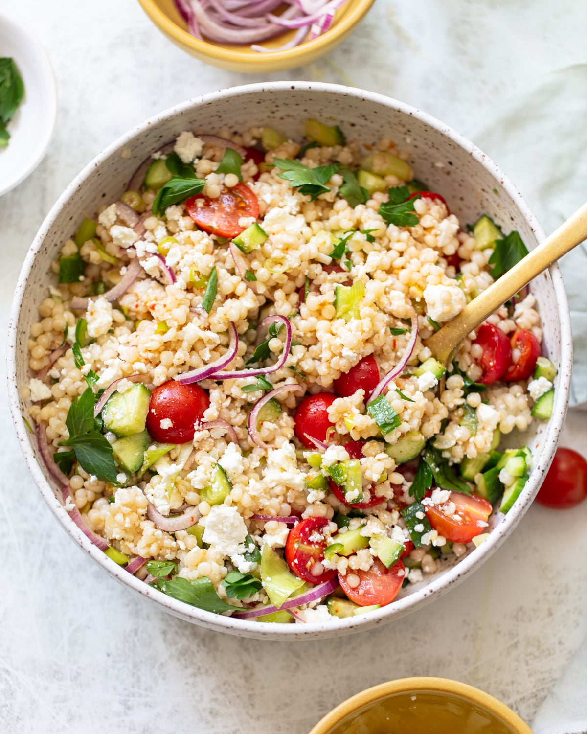 A bowl of couscous salad with cherry tomatoes, sliced red onions, cucumber, feta cheese, and fresh parsley, served with a wooden spoon on a light-colored table.