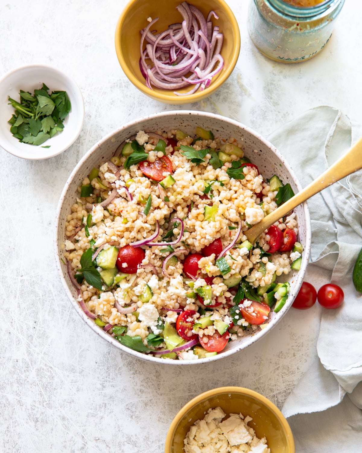 A bowl of couscous salad with cherry tomatoes, cucumbers, parsley, and red onions, surrounded by small bowls of sliced red onion, parsley, and feta cheese on a white surface.