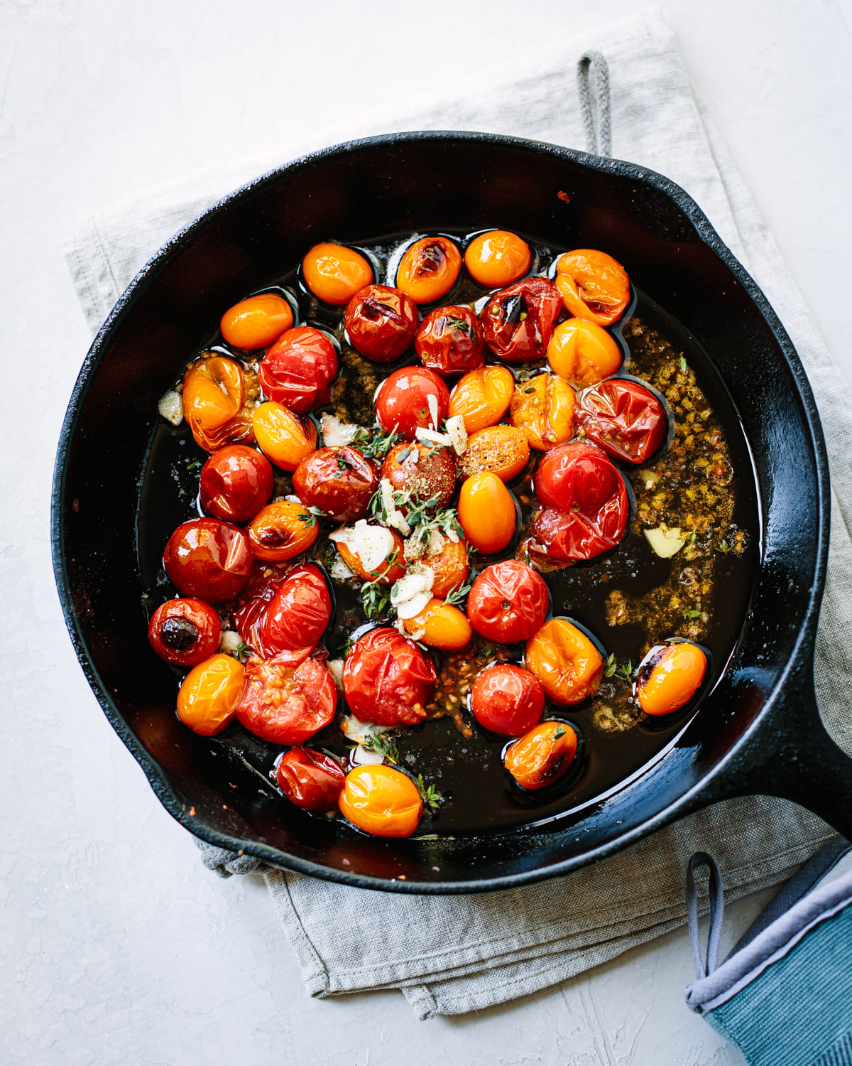 A black cast-iron skillet with roasted red and yellow cherry tomatoes, garlic, herbs, and oil sits on a light cloth and white surface.