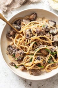 A bowl of linguine with creamy sauce, sautéed mushrooms, grated parmesan, and fresh herbs, with a fork twirled into the pasta.