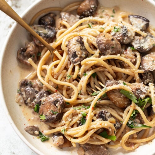 A bowl of linguine with creamy sauce, sautéed mushrooms, grated parmesan, and fresh herbs, with a fork twirled into the pasta.