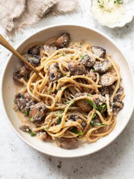A bowl of pasta with creamy mushroom sauce, garnished with chopped herbs and grated cheese, with a fork twirling some pasta. A beige cloth and a small dish of cheese are in the background.