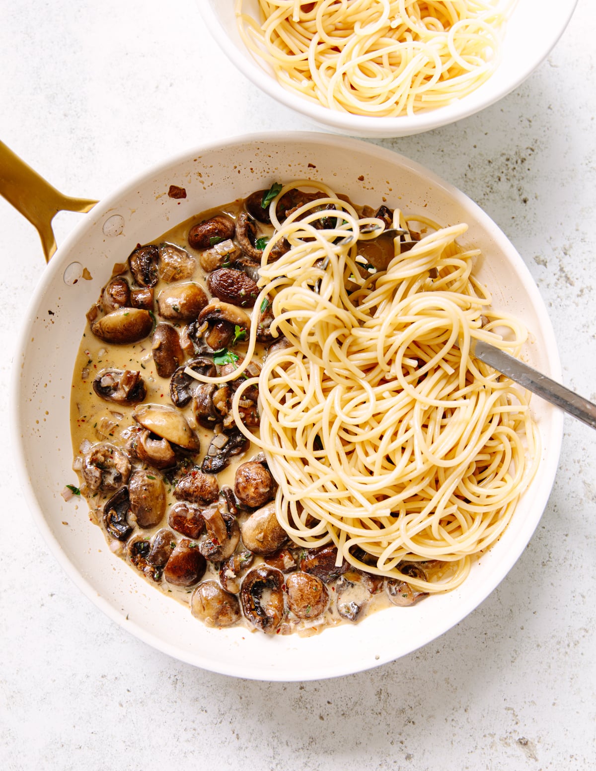A white saute pan containing cooked linguine pasta and creamy sautéed mushrooms sits next to a bowl of plain pasta on a light surface. A fork rests in the pan, ready to mix the pasta with the mushroom sauce.