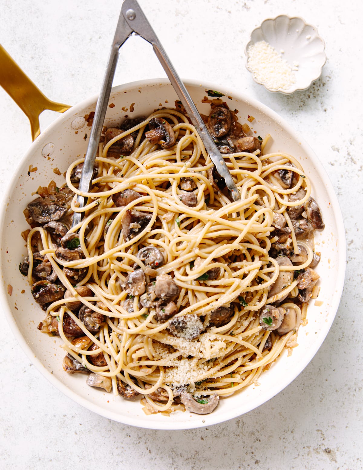 A cream colored skillet filled with spaghetti, sautéed mushrooms, and herbs, topped with grated cheese, with metal tongs resting inside. A small dish of grated cheese is nearby on a light-colored surface.