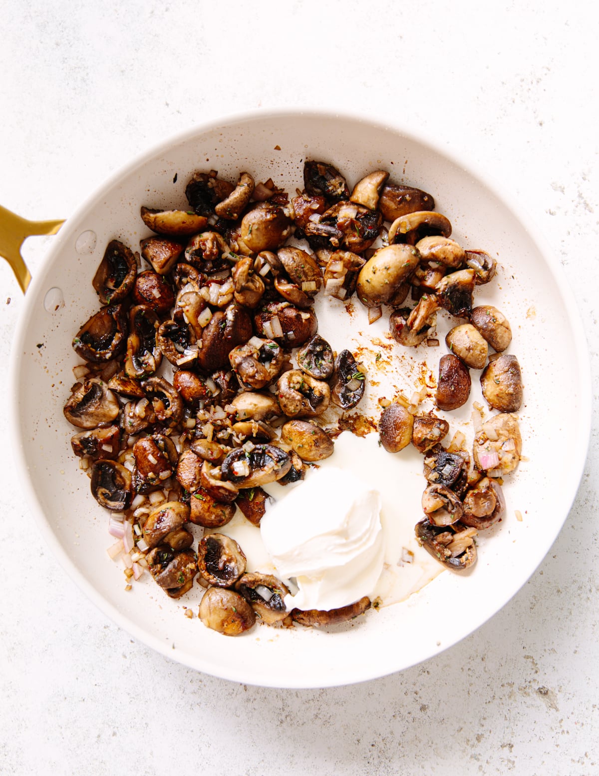 A white skillet containing sautéed mushrooms and onions with a dollop of creme fraiche in the center, resting on a white surface.