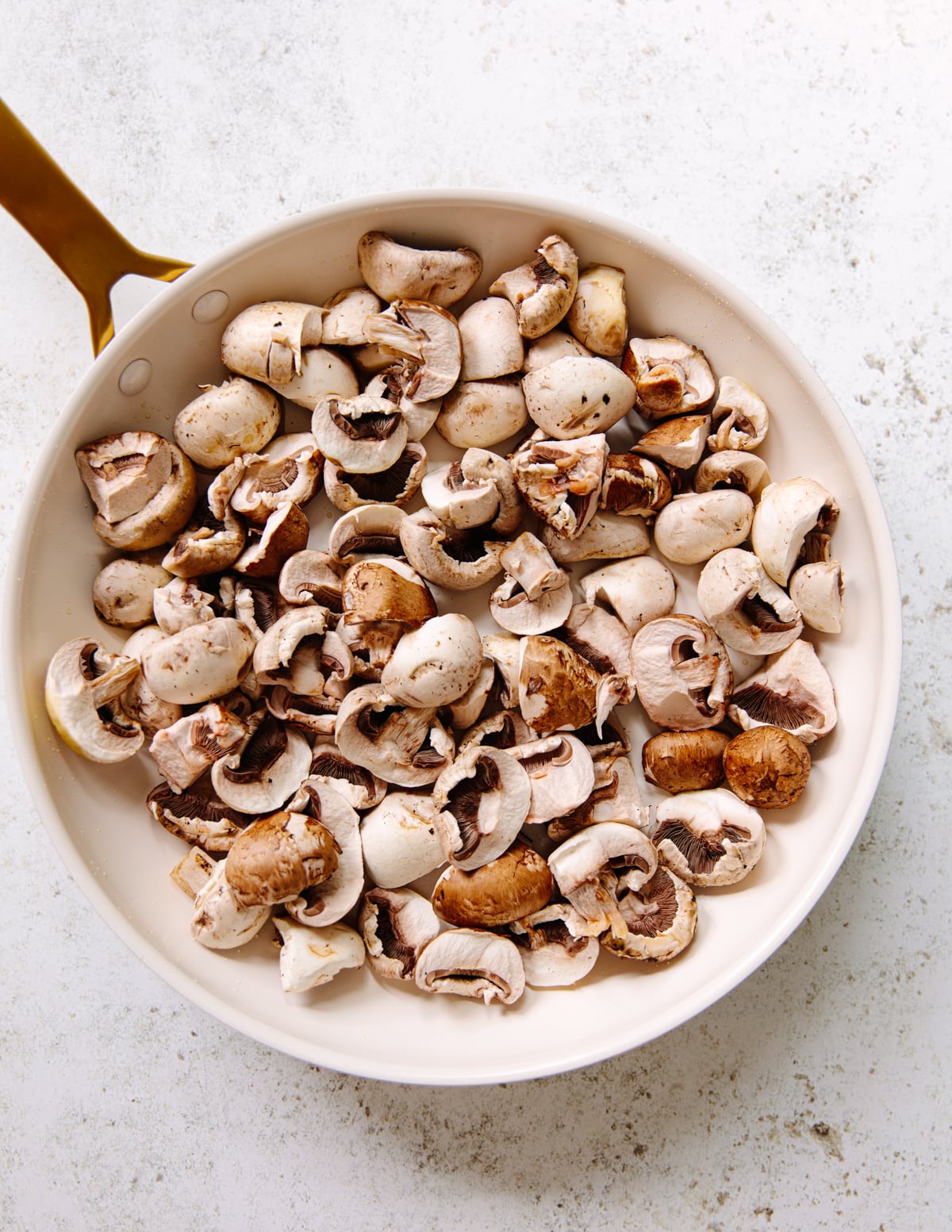A wide skillet filled with sliced brown and white mushrooms, placed on a light-colored surface. The pan has a gold-colored handle.