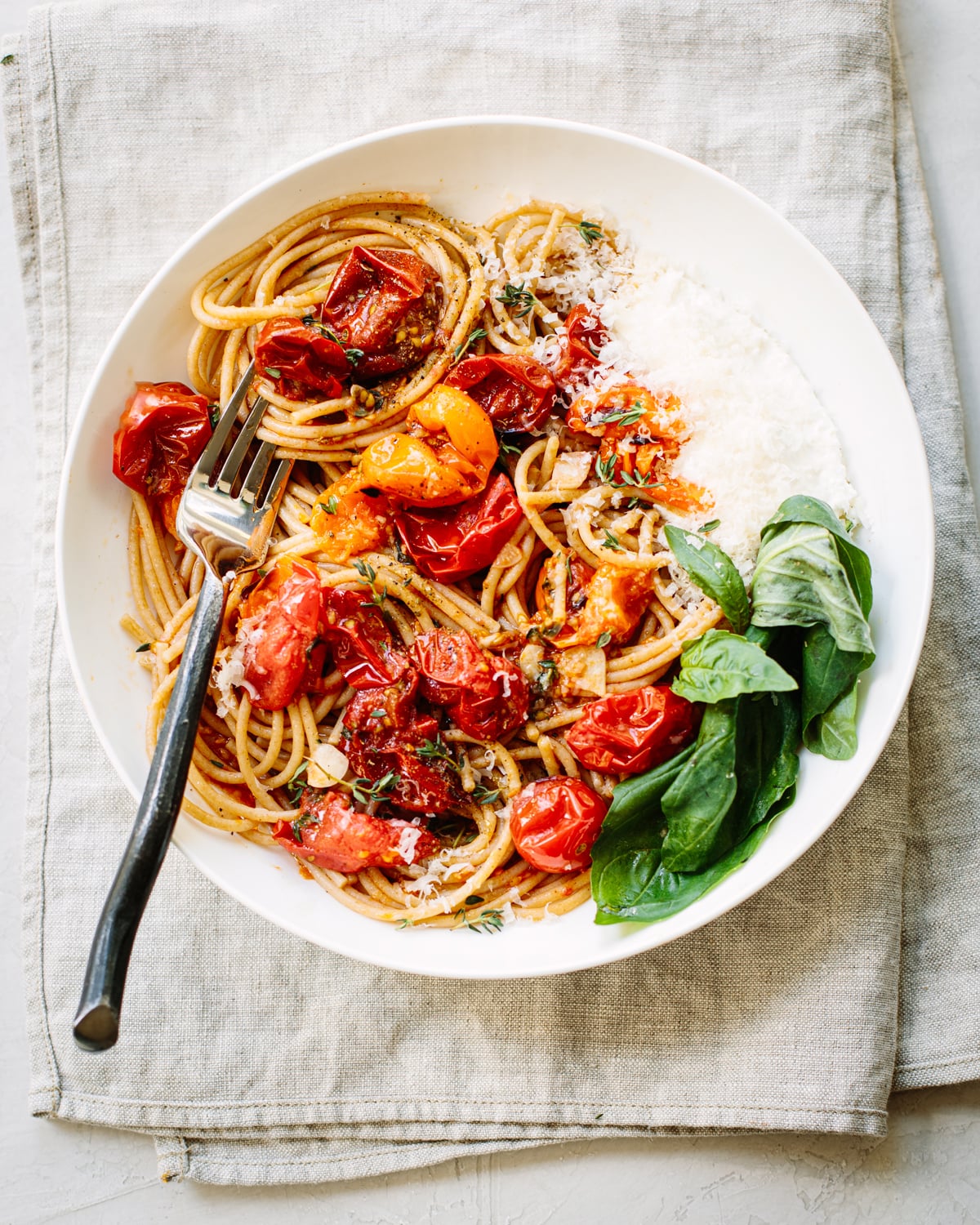 A white plate of spaghetti with roasted cherry tomatoes, grated cheese, fresh basil leaves, and a fork, placed on a folded beige cloth napkin.
