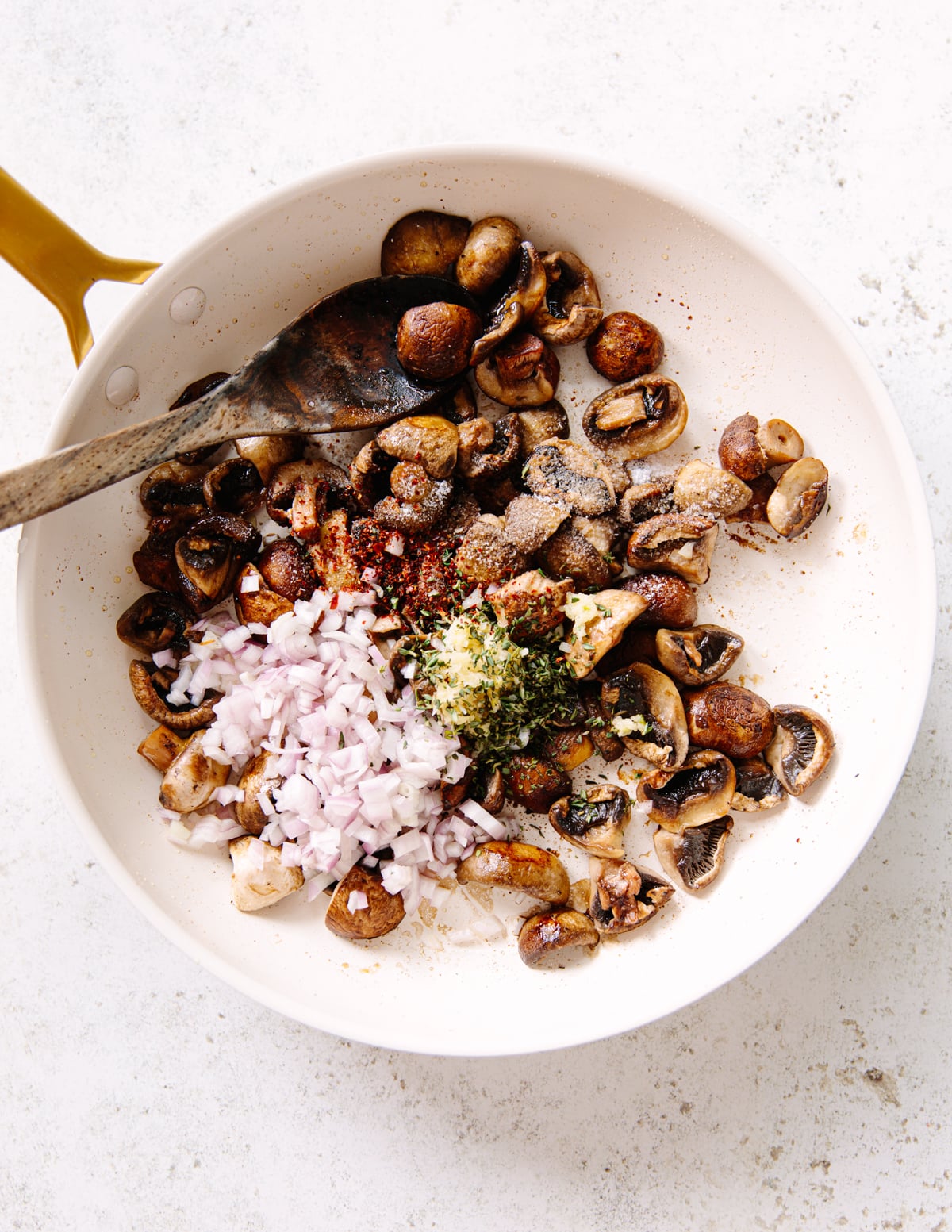 A white frying pan with chopped mushrooms, minced garlic, diced shallots, fresh herbs, and spices being stirred with a wooden spoon on a light countertop.