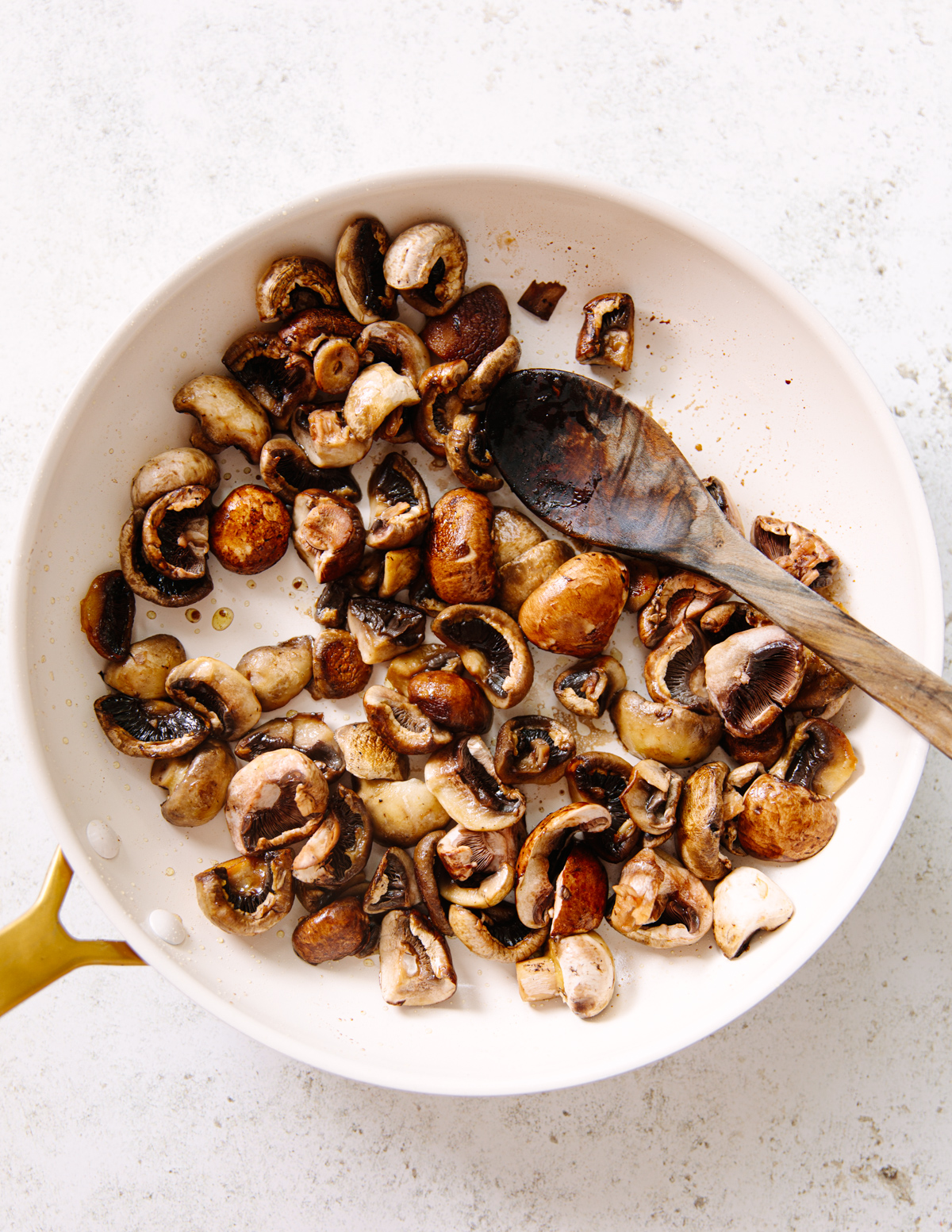 Sliced mushrooms being sautéed in a white frying pan with a wooden spoon resting on the side, on a light-colored surface.